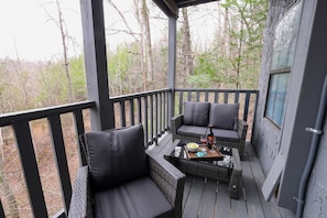 A covered outdoor seating area with wicker furniture and cushions, a table with a bottle and glasses, and a view of a forested area. - Starlight Summit Big Buck Hunter Arcade Hot Tub (Sevierville)