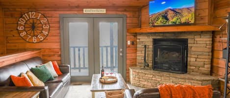 Interior acogedor de una cabaña con chimenea, un televisor montado encima, un sofá de cuero con cojines y un reloj de pared de madera.