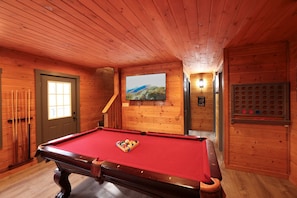 Une table de billard se trouve dans une pièce en bois, équipée d’une porte, d’une fenêtre et d’un téléviseur fixé au mur.