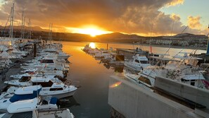 Un port de plaisance où sont amarrés de nombreux bateaux, un coucher de soleil en arrière-plan et une vue sur la ville qui s’étend de l’autre côté de l’eau.