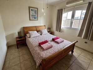 Un dormitorio con una cama de madera, dos mesitas de noche, una lámpara de noche, una ventana con cortinas y un aire acondicionado de pared.
