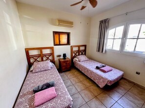 Une chambre avec deux lits, une table de chevet, un ventilateur de plafond, un appareil de climatisation, une fenêtre avec des rideaux et un tableau encadré au mur.
