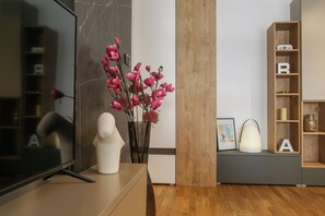 A modern living room with a flat-screen TV, a white bust sculpture, a vase with pink flowers, and a wooden bookshelf with a framed picture. - Supernova DeLuxe Residence 2bdr Apartment in Novi Sad with AC for relaxing stays (Novi Sad)