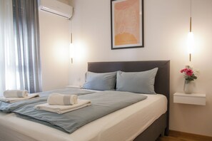 A neatly made bed with pillows and folded towels, a bedside table with a vase of flowers, and a wall-mounted air conditioner. - Supernova DeLuxe Residence 2bdr Apartment in Novi Sad with AC for relaxing stays (Novi Sad)