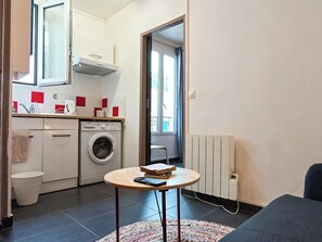 A compact living space with a washing machine, a radiator, a small table with books, and a view through an open door. - New Listing (Paris)