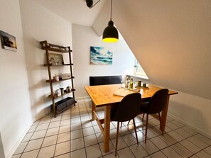 A dining area with a wooden table, two chairs, a shelf with kitchen items, a painting on the wall, and a pendant light. - Island Bliss Fohr (Wrixum)