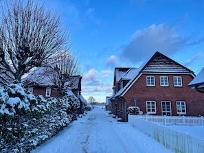 A snow-covered residential street with houses on both sides, a white picket fence, and a clear blue sky. - Island Bliss Fohr (Wrixum)