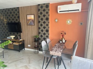 Un saló modern amb un televisor de pantalla plana, una taula de dinar amb cadires, una taula auxiliar petita amb una planta, i aire condicionat fixat a la paret.
