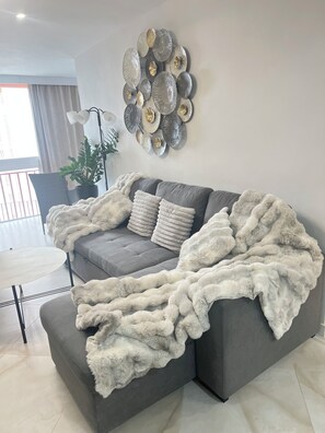 Un saló modern amb un sofà seccional gris, una manta blanca i esponjosa, i una peça decorativa a la paret.