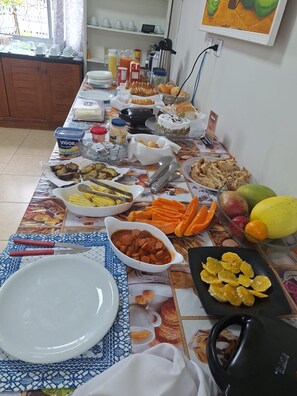 Una mesa de comedor con diversos alimentos y utensilios, una zona de cocina con armarios y un ventanal, y un enchufe en la pared.