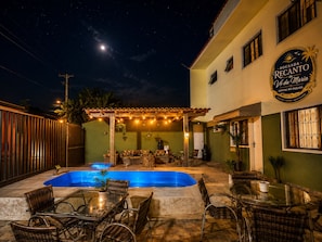 Un patio exterior nocturno con piscina, mobiliario de mimbre y una zona de estar cubierta.