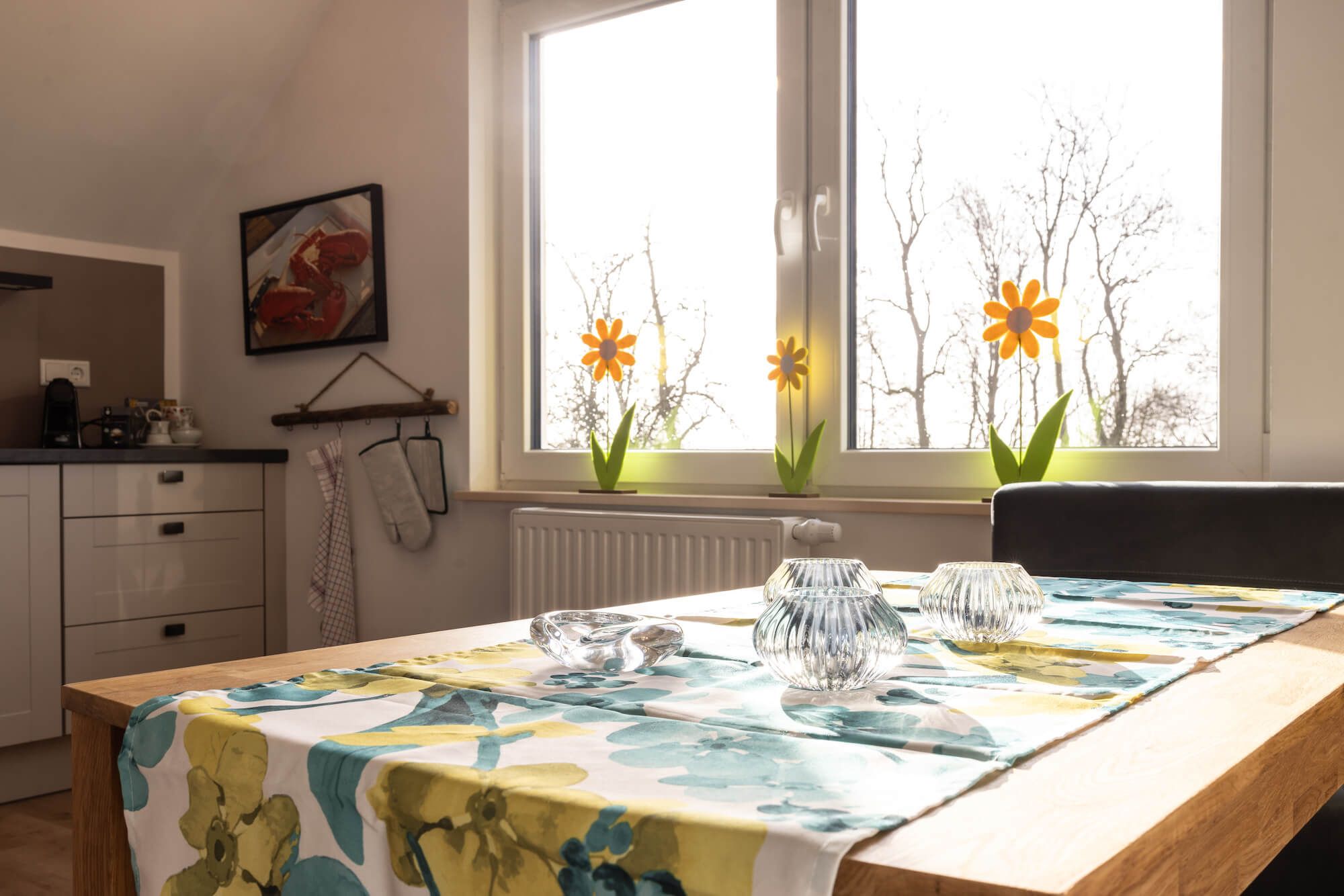 Ein Essbereich mit einem Tisch für zwei Personen, einem Fenster mit Blumen und einer Küche im Hintergrund.