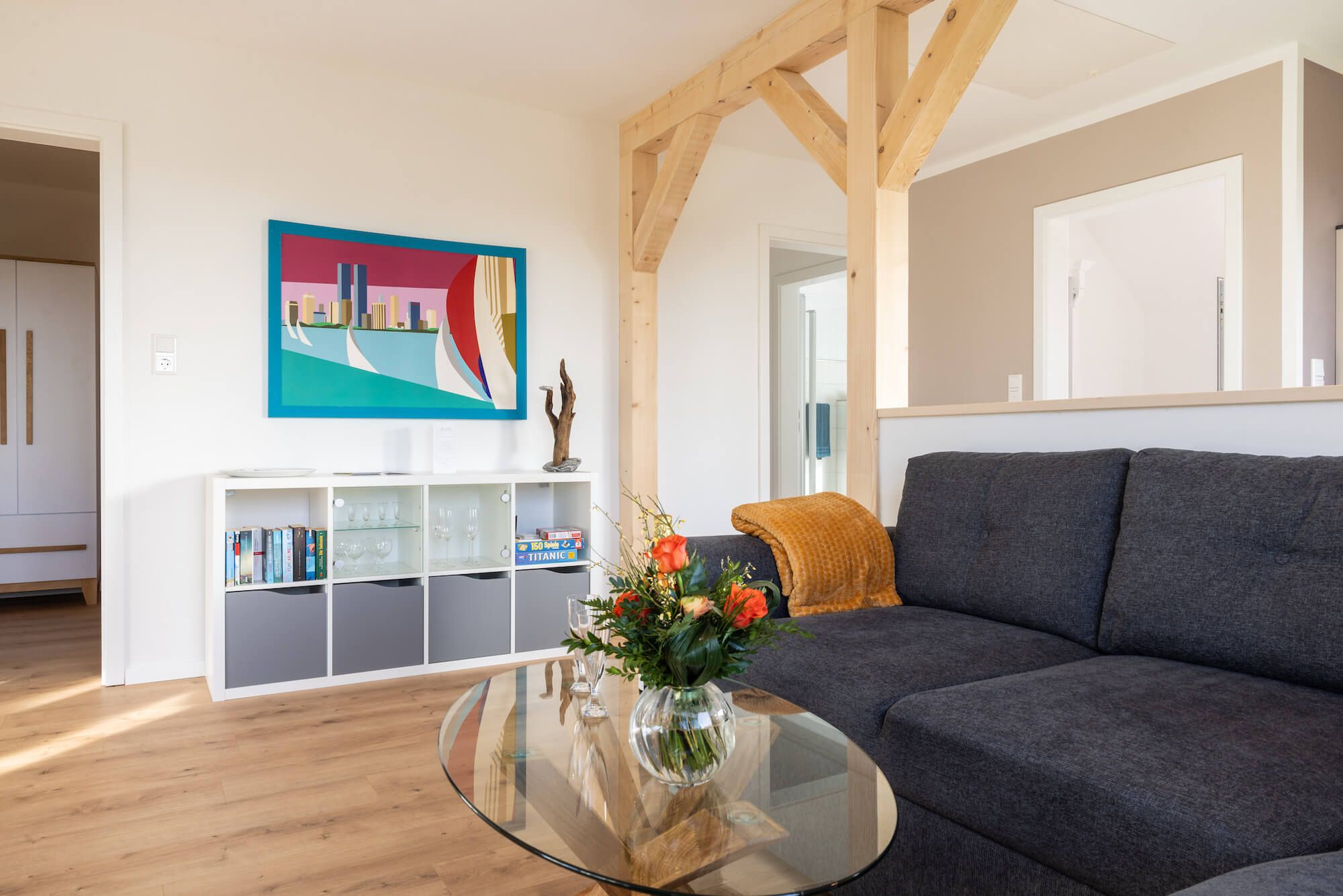 Ein Wohnzimmer mit einem dunkelgrauen Sofa, einem Glastisch mit Blumen, einem Bücherregal mit Büchern und Dekorationsobjekten sowie einem farbenfrohen Bild an der Wand.