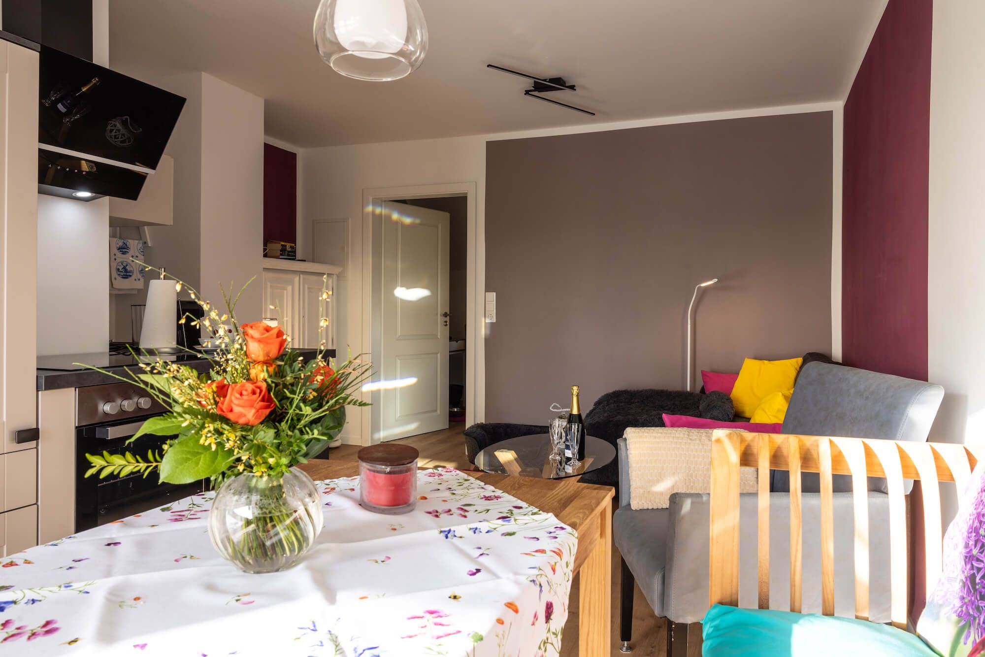 Een moderne woonkamer met een eethoek, een bank met kleurrijke kussens, een vaas met bloemen op de eettafel, en een flatscreen televisie aan de muur.