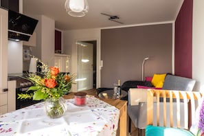 Een moderne woonkamer met een eethoek, een bank met kleurrijke kussens, een vaas met bloemen op de eettafel, en een flatscreen televisie aan de muur.