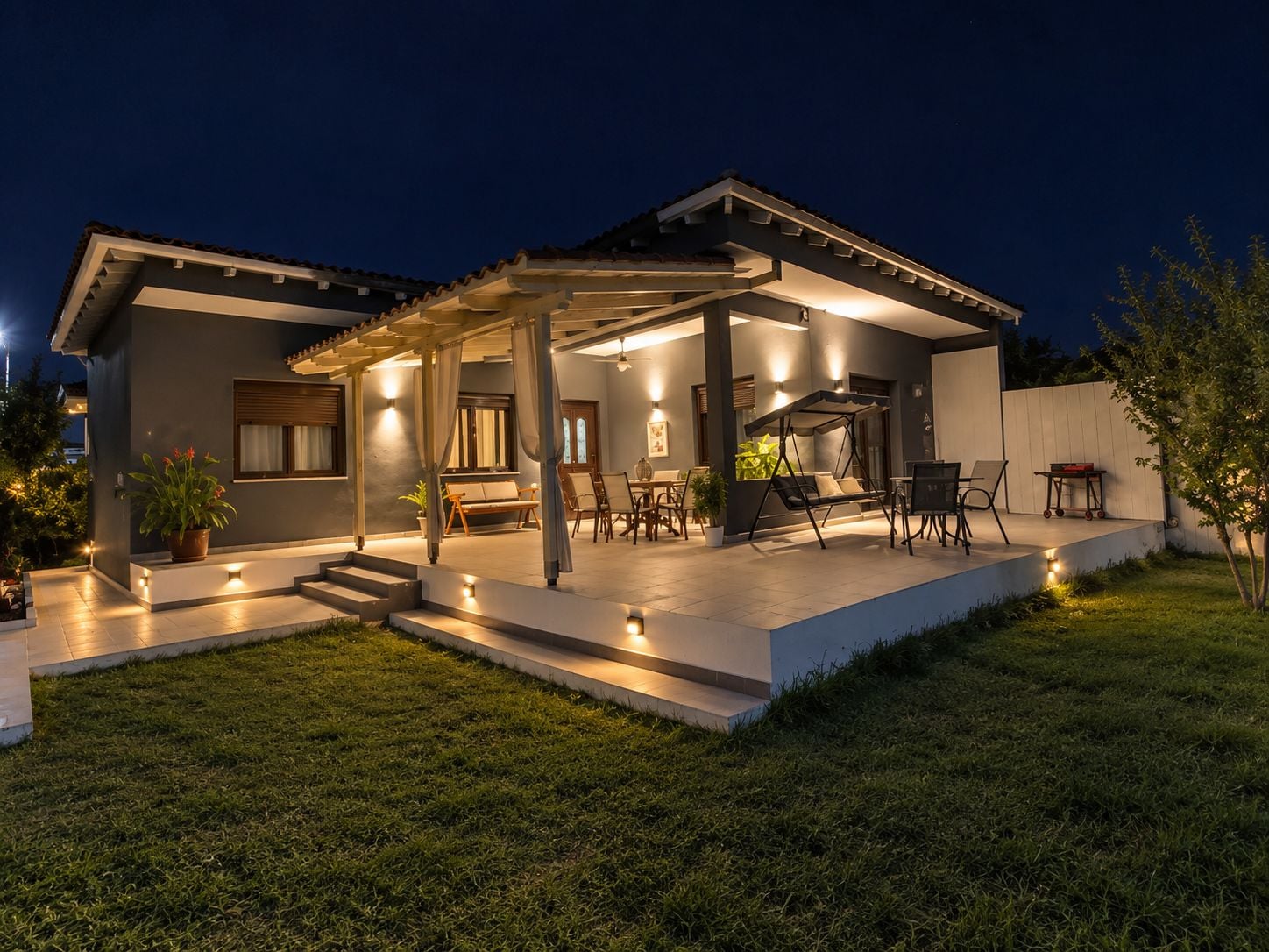 Una casa moderna con porche cubierto, mobiliario exterior y un jardín bien iluminado.