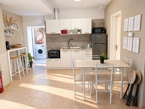 Una cocina moderna con armarios blancos, un refrigerador de acero inoxidable, un microondas y un lavamanos. Hay una mesa de comedor con sillas y una planta en maceta.