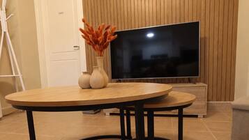 Ett modernt vardagsrum med en väggpanel i trä, en stor platt-TV, ett runt soffbord med ben i svart metall och en vas med torkade blommor.