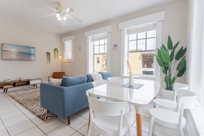 A modern living room with a blue sofa, white dining table, and chairs, a ceiling fan, and large windows. - Historic River District Downtown Escape (Fort Myers)
