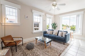 A living room with a blue sofa, a glass coffee table, a brown leather chair, a patterned rug, and large windows with blinds. - Historic River District Downtown Escape (Fort Myers)