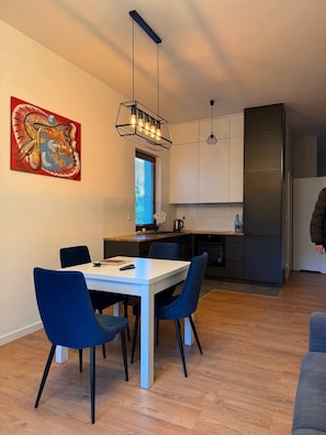 Une cuisine moderne dotée d’un réfrigérateur encastré, un coin repas avec une table blanche et des chaises bleues, et une peinture abstraite aux couleurs vives accrochée au mur.