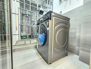 A compact, modern washing machine with a front-loading door, situated in a clean, tiled room with a visible laundry basket. - Cozy 1-bedroom apartment in awesome Nairobi with gym and pool- Kilimani (Nairobi)