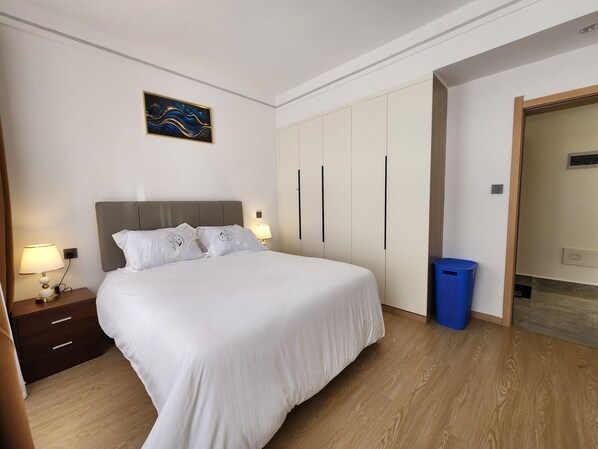 A hotel room with a bed, nightstands, a wall-mounted artwork, a wardrobe, and a trash bin. - Cozy 1-bedroom apartment in awesome Nairobi with gym and pool- Kilimani (Nairobi)