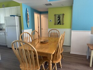 A dining area with a wooden table and chairs, a kitchen with white cabinets, and a blue wall with a lighthouse painting. - Siesta Sunset Royale #12 3 Bedroom Apts (Siesta Key)