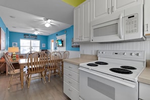 A kitchen with white cabinets, a microwave, an oven, and a dining area with wooden chairs and a table. - Siesta Sunset Royale #456 4 Bedroom Apts (Siesta Key)
