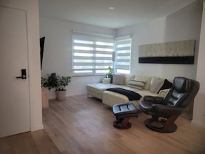 Ein modernes Wohnzimmer mit einer Couch, einem schwarzen Ledersessel, einer Topfpflanze und einem großen Fenster mit Jalousien.