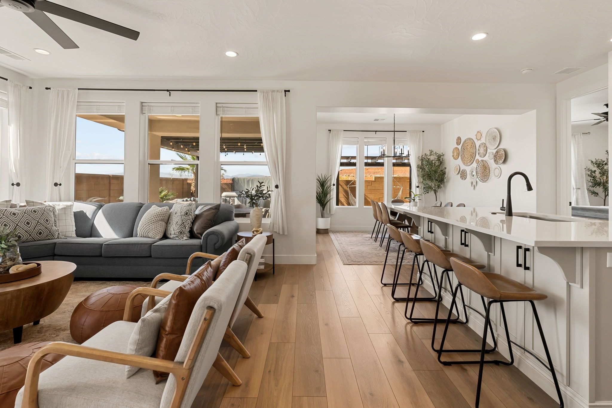 A modern living room with a grey sofa, wooden coffee table, and leather armchairs. A kitchen area with bar stools and a white countertop is visible.