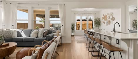 A modern living room with a grey sofa, wooden coffee table, and leather armchairs. A kitchen area with bar stools and a white countertop is visible.