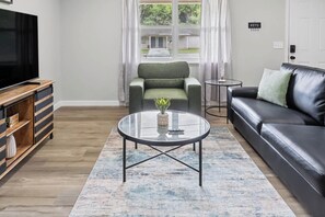Ein modernes Wohnzimmer mit Fernseher, einem grünen Sessel, einer schwarzen Ledersofa und einem Glastisch.
