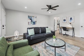 Ein modernes Wohnzimmer mit einem schwarzen Ledersofa, einem Glastisch und einem grünen Sessel. Ein Deckenventilator ist vorhanden, im Hintergrund ist eine Küchenzeile zu sehen und die Wände sind mit abstrakter Kunst dekoriert.