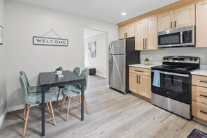 Eine moderne Küche mit einem Edelstahl-Kühlschrank, einer Mikrowelle und einem Backofen. Ein Esstisch mit Stühlen und ein 'WELCOME'-Schild an der Wand.