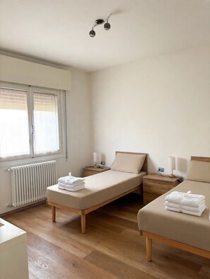 A minimalist bedroom with two beds, a radiator, a bedside table with a lamp, and a window with blinds. - New Listing (Noale)