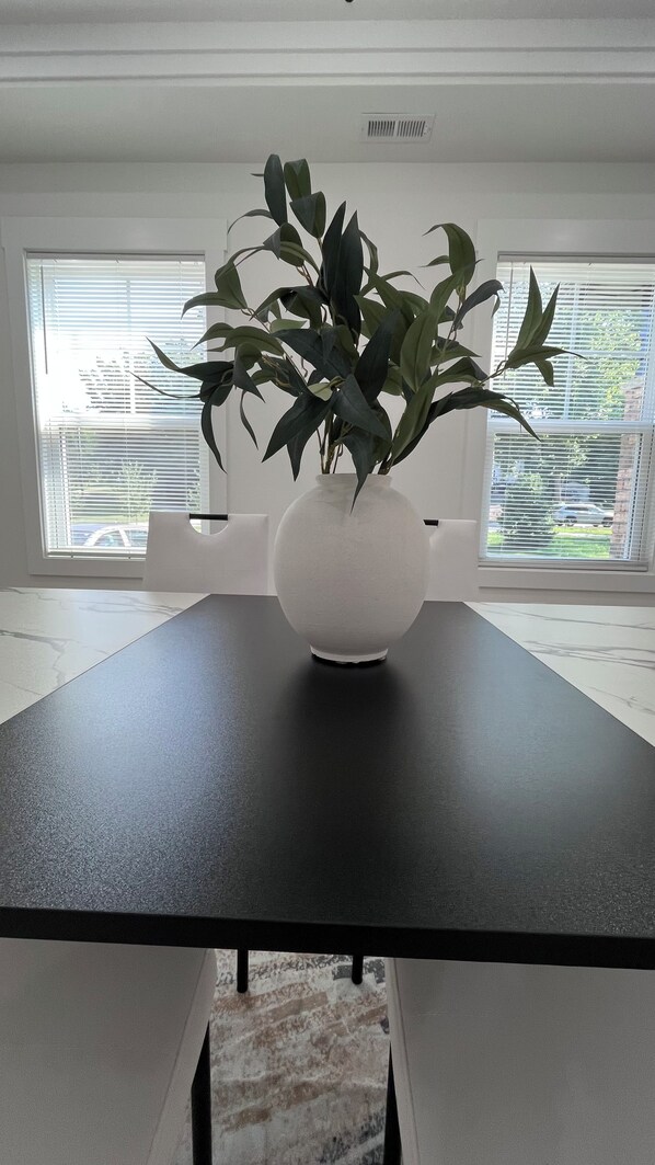 A modern room with a black table, a white vase with green plants, and large windows with blinds. - The Best Place for Family Stays, and Small Group (Mint Hill)