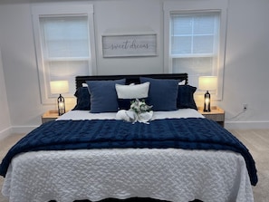 A bedroom with a bed, two bedside lamps, a wall-mounted sign, and a window with blinds.