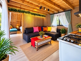 Ein gemütliches Blockhaushaus-Interieur mit Holzbalkendecke, einer Couch mit bunten Kissen, einer Küchenzeile und einem Esstisch.