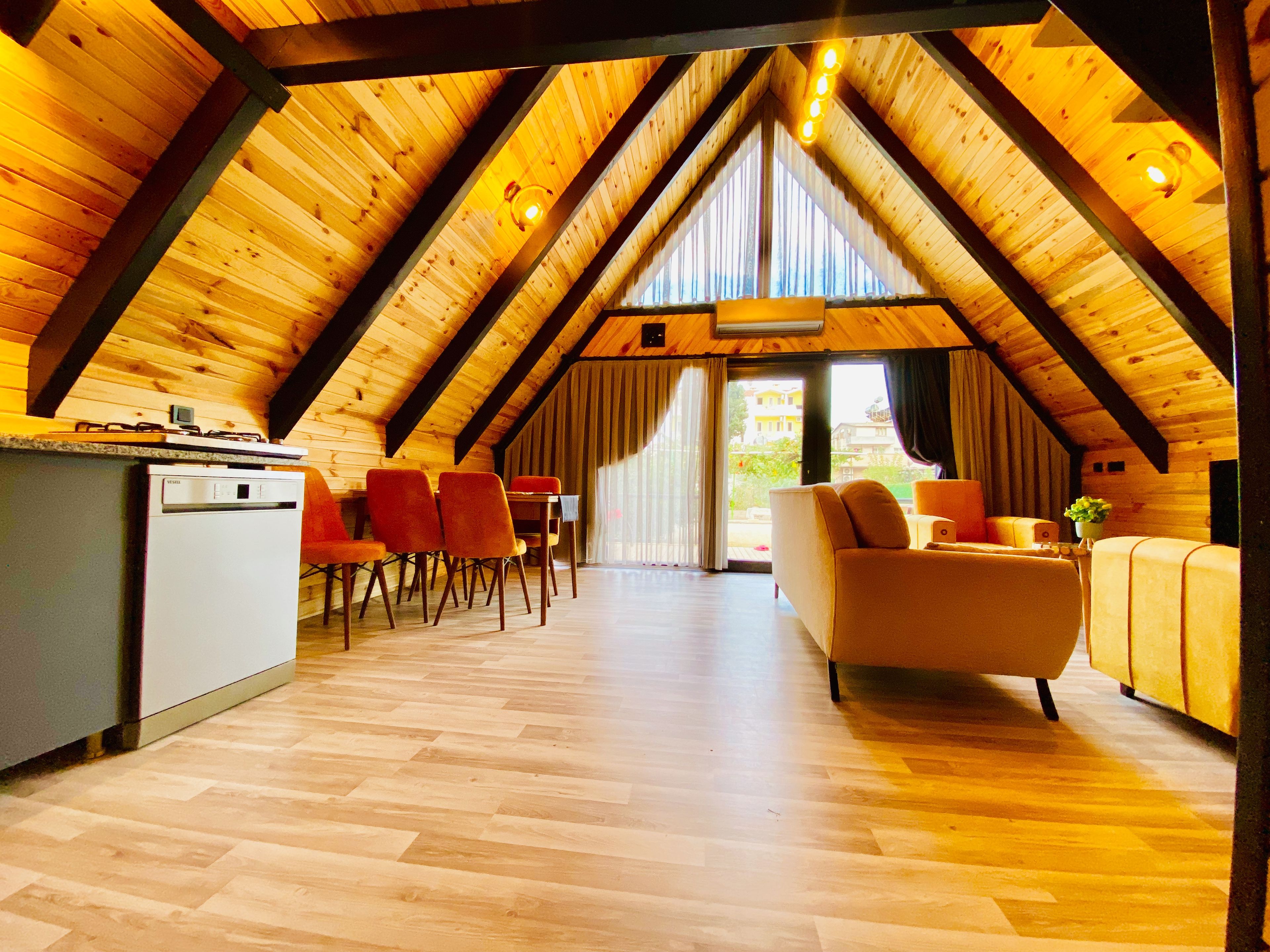 A spacious attic room with a wooden ceiling, a dining area with chairs, a sofa, and a kitchen area with a refrigerator.