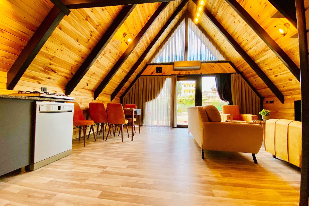Un'ampia stanza mansardata con soffitto in legno, zona pranzo con sedie, un divano e angolo cottura con frigorifero.