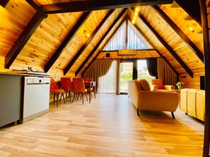 A spacious attic room with a wooden ceiling, a dining area with chairs, a sofa, and a kitchen area with a refrigerator. - Village Garden (Antalya)