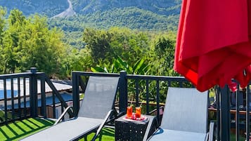 A balcony with two lounge chairs and a red umbrella, overlooking a mountainous landscape.