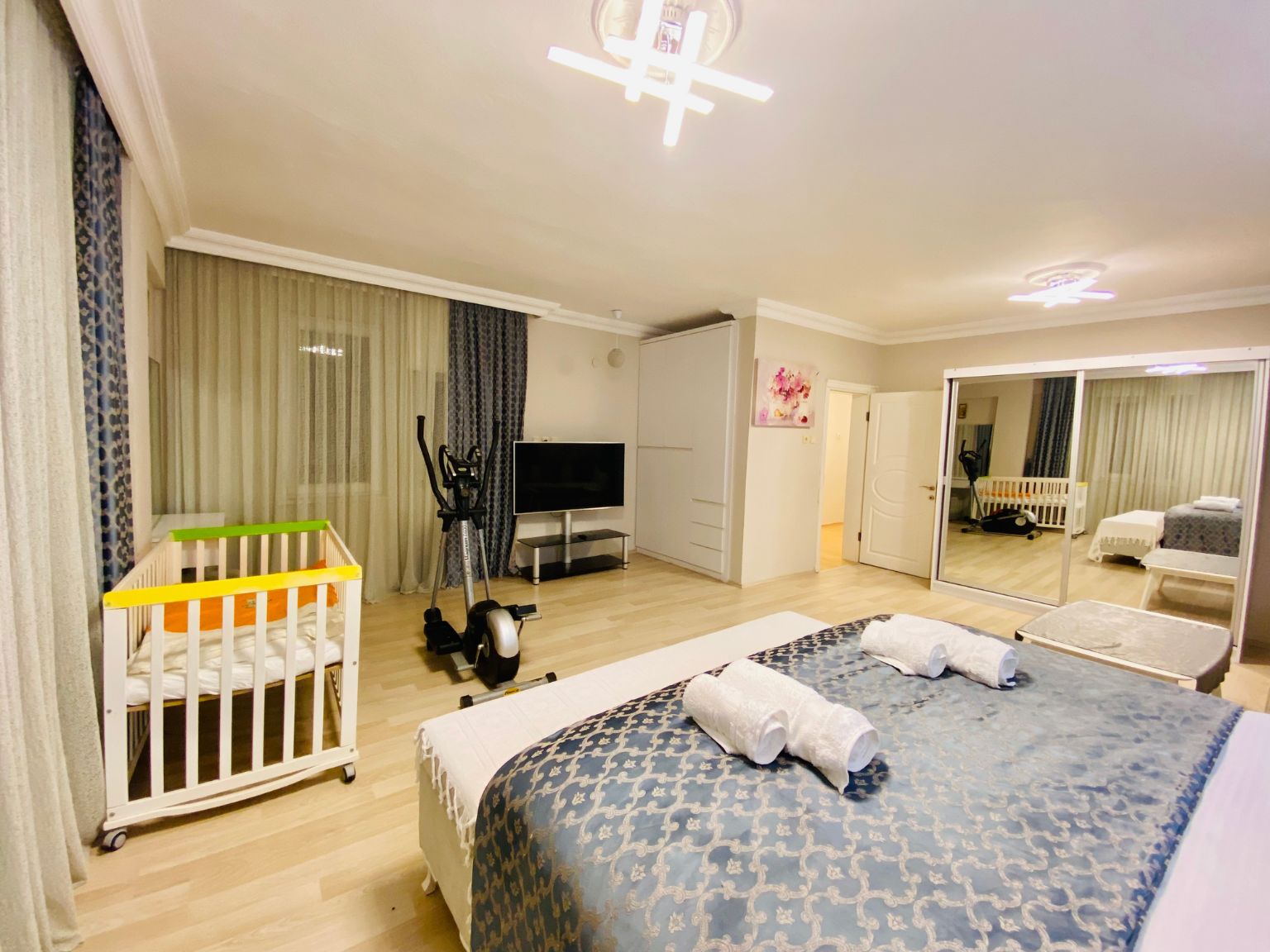 Une chambre d’hôtel comprenant un lit, un berceau, une télévision, un miroir et une porte donnant sur une autre pièce.