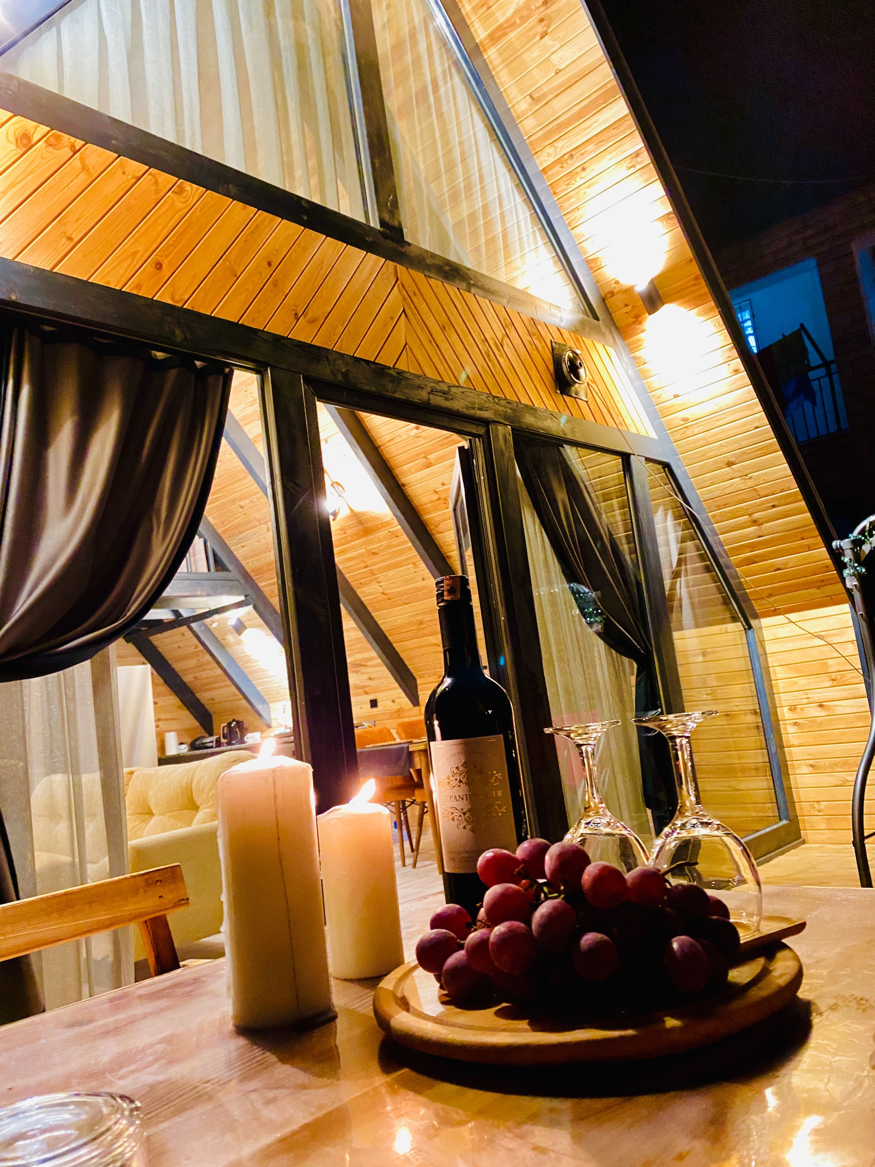 Une table avec une bouteille de vin, des bougies et un assortiment de fruits, dans une pièce aux murs en bois et aux grandes fenêtres.