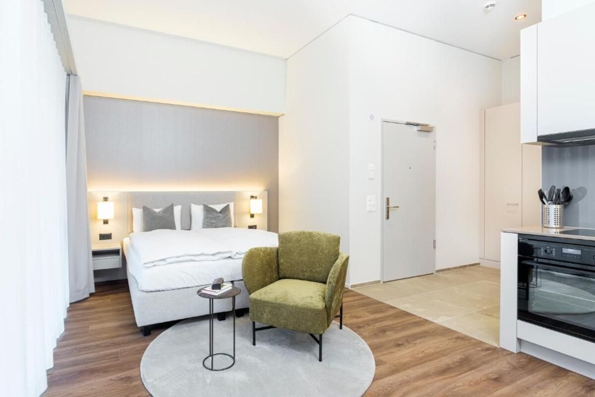 A modern hotel room with a bed, a small round table, a green armchair, a kitchen area with a stove, and a door leading to another room.