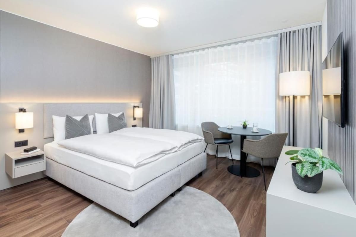 A modern hotel room with a large bed, bedside tables, a desk with chairs, a round table, and a potted plant.