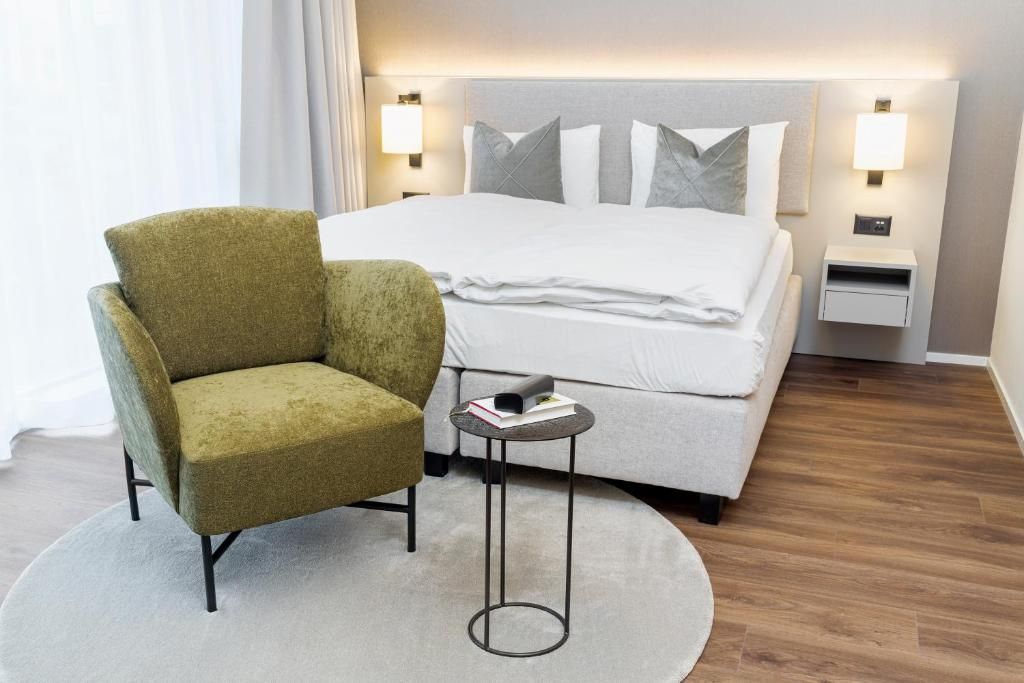 A hotel room with a bed, a green armchair, a small round table, and a nightstand.