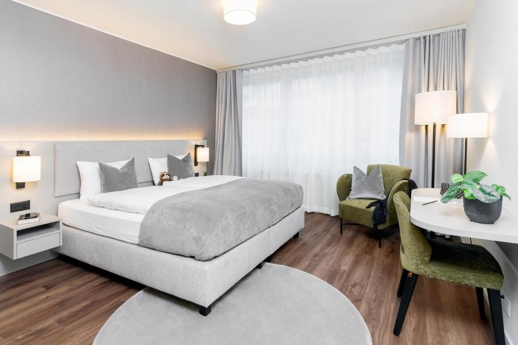 Camera d'hotel moderna con un letto grande, lampade ai comodini, una poltrona verde, un piccolo tavolino con una pianta e vista all'esterno tramite tende trasparenti.