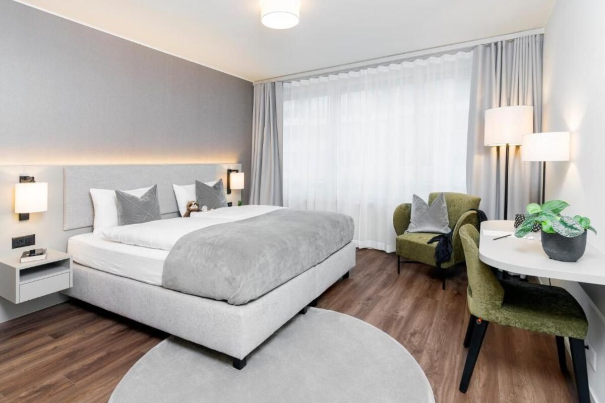 A modern hotel room with a large bed, bedside lamps, a green armchair, a small table with a plant, and a view of the outside through sheer curtains.